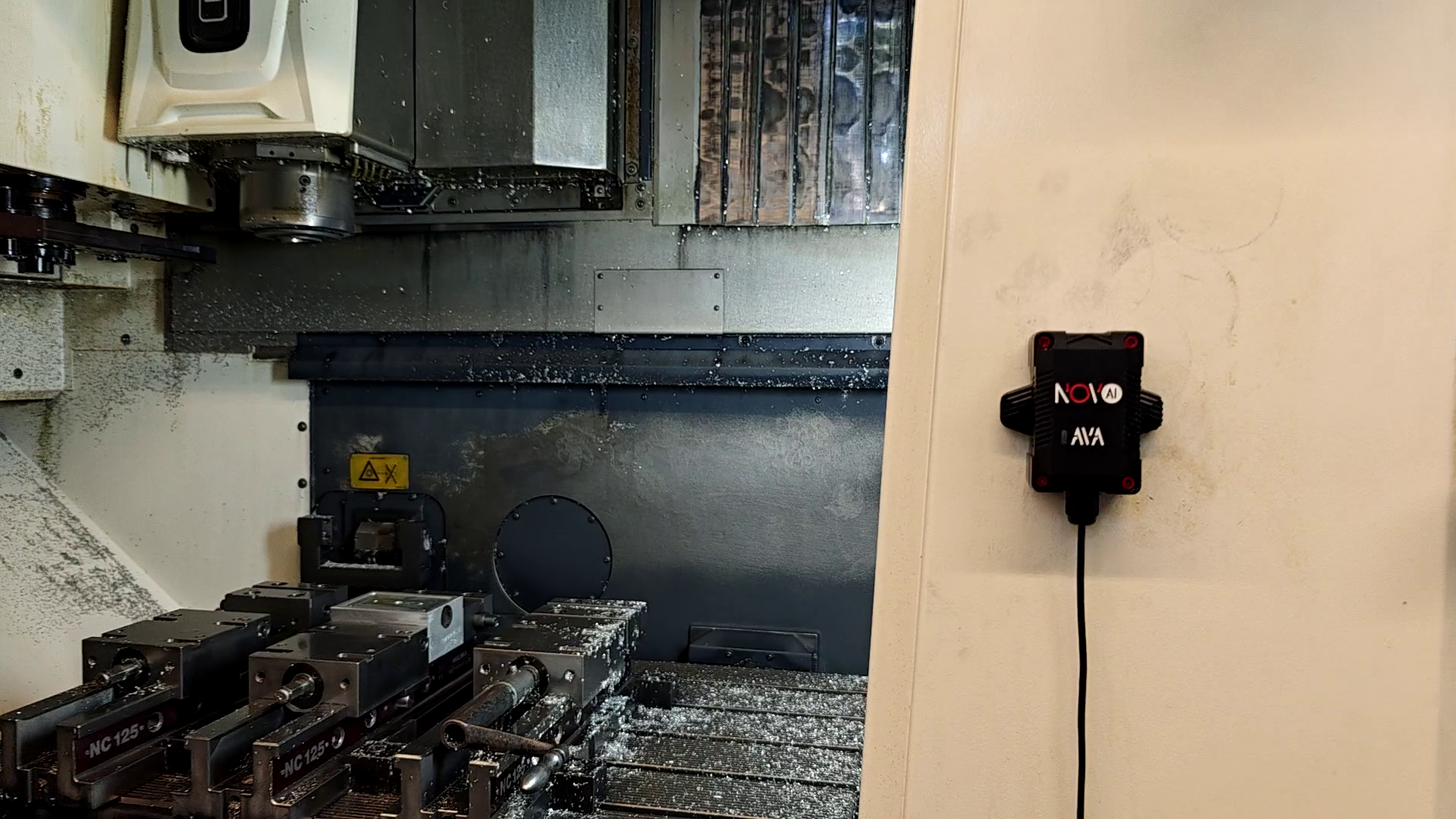Schwarzer Novo AI AVA-Sensor am hellen Geh&auml;use einer ge&ouml;ffneten CNC-Fr&auml;smaschine mit sichtbaren Schraubst&ouml;cken und Sp&auml;nen.